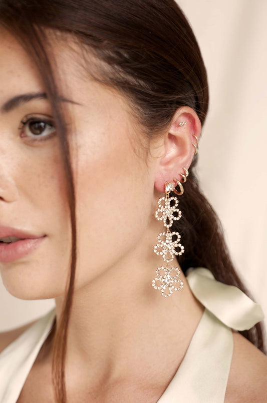 A woman's ear adorned with elegant, sparkling flower-shaped earrings made of intricate designs, set against a softly blurred, neutral background, highlighting her subtle features and glowing skin.