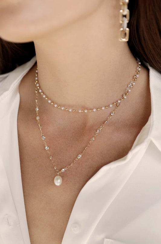 A layered gold necklace featuring delicate pearls and crystals sits elegantly against the skin, complemented by a white button-up shirt in a soft-lit, neutral background.