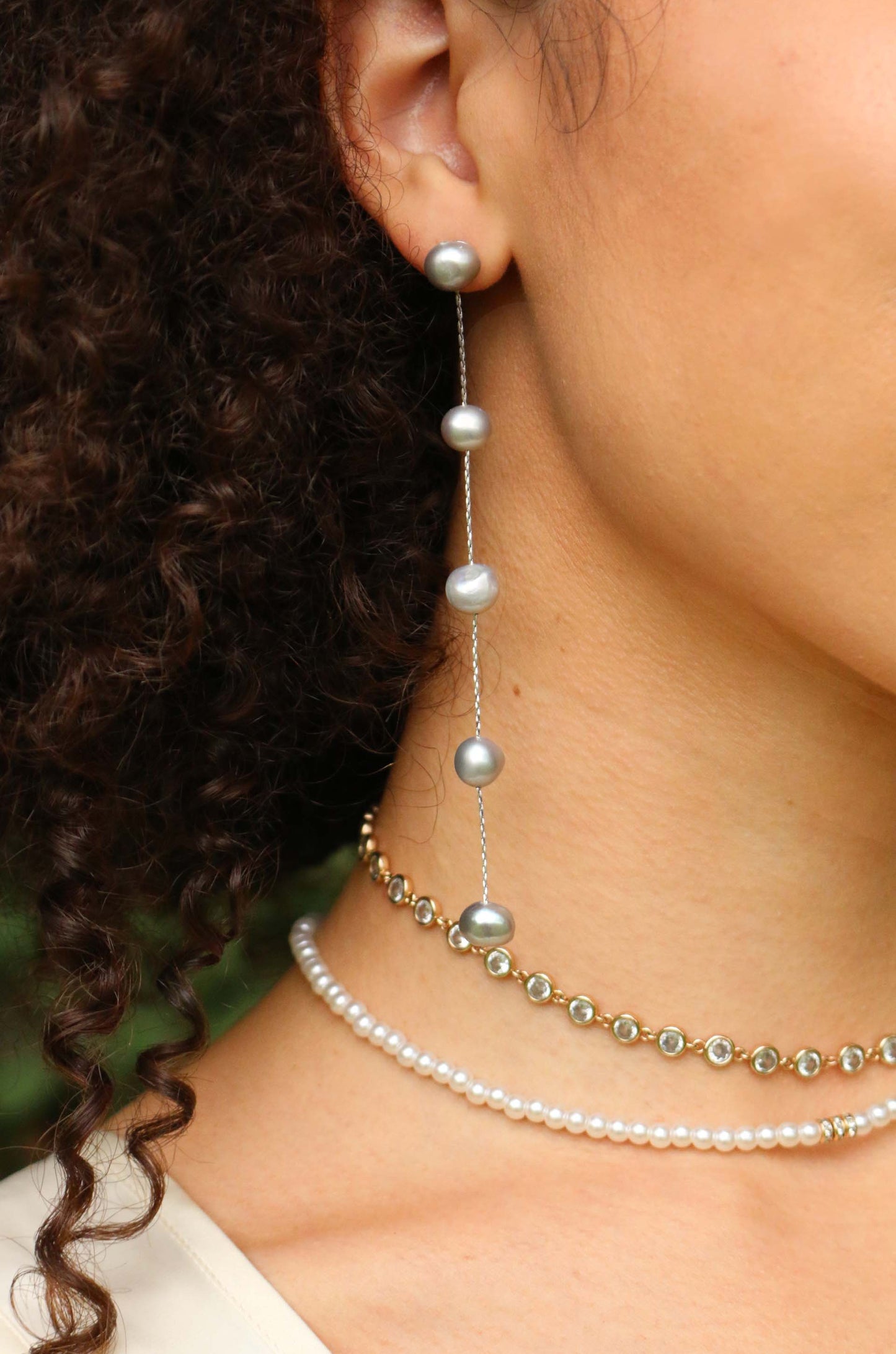 A person wears long pearl earrings and layered pearl and gold chokers, showcasing elegant jewelry against a blurred green background, reflecting a natural and fashionable style.