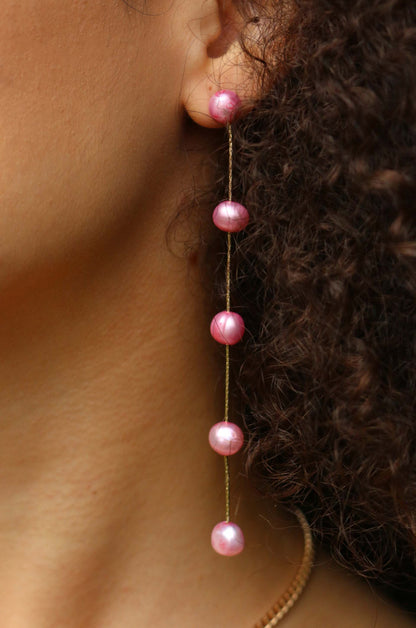 A single earring featuring four pink pearls hangs from a woman's ear. Her curly hair frames the earring against her skin, highlighting the delicate design of the jewelry.