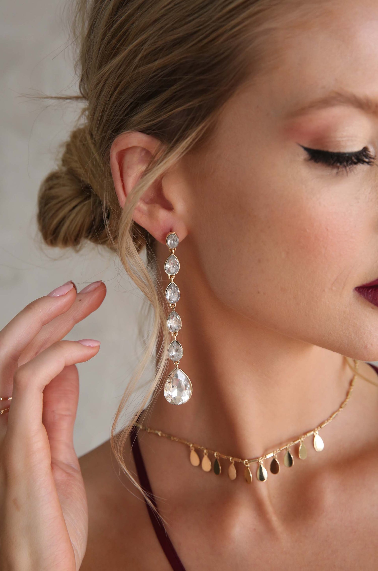 A woman's ear features a long, sparkling earring composed of crystal drops. She gently touches her neck, adorned with a gold choker, set against a soft, neutral background.