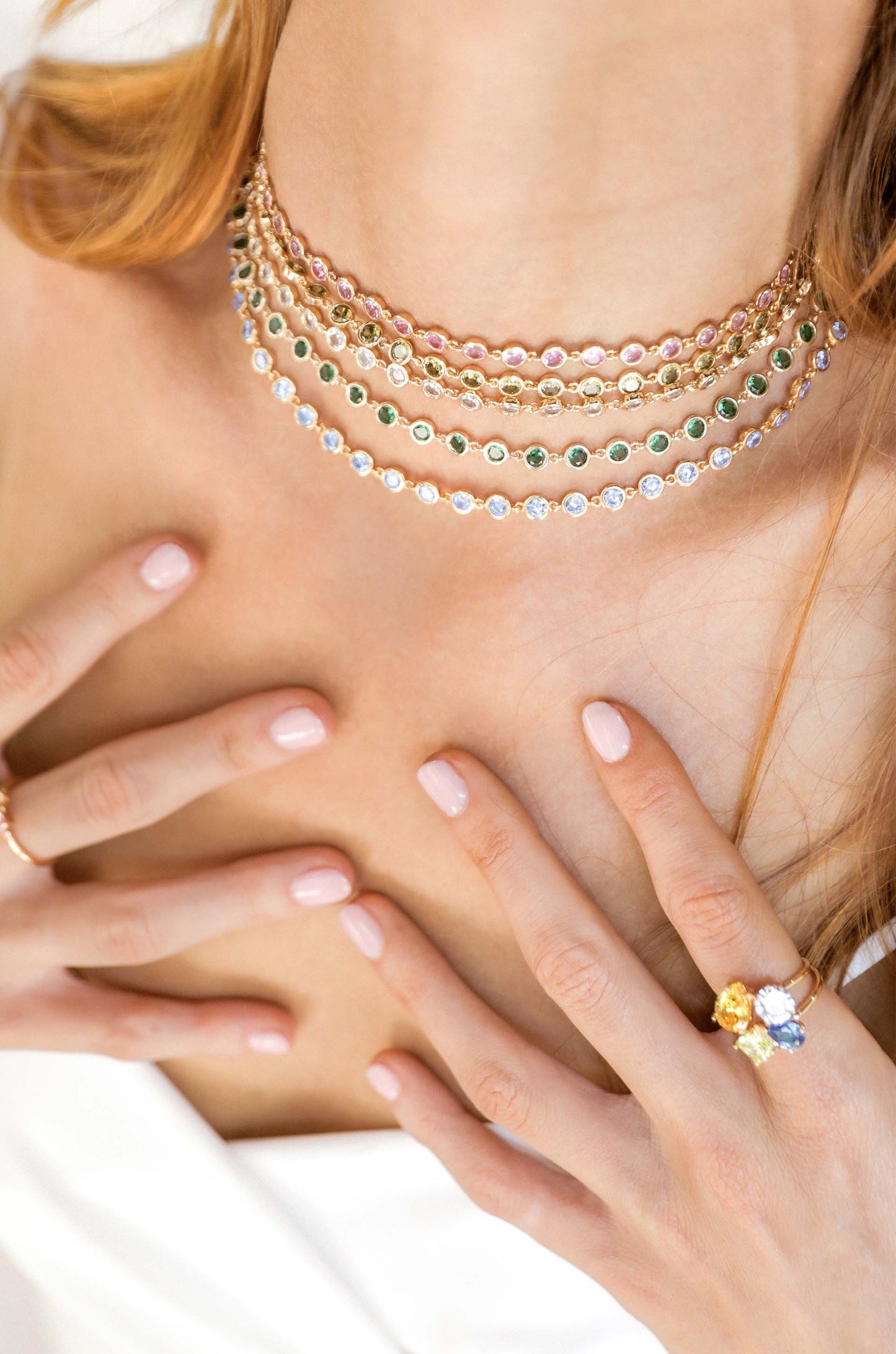 A multi-layered gemstone necklace adorns a person's neck while two hands with polished nails gently touch the area, set against a light background, suggesting a soft, elegant atmosphere.