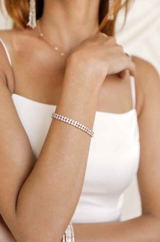 A hand adorned with a sparkling bracelet rests gently on a shoulder, set against a soft, neutral background. The person is wearing a simple white top, enhancing the elegant jewelry.