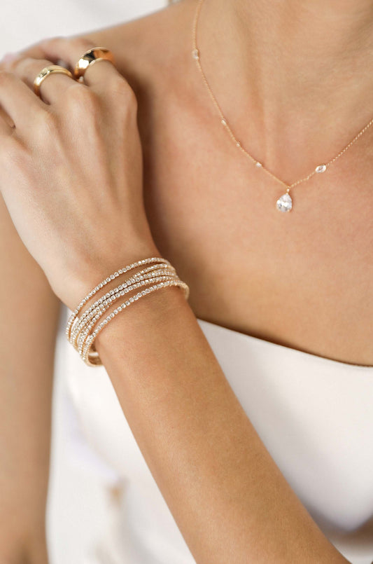 A hand adorned with multiple glittering bracelets and rings gracefully touches a shoulder. A delicate necklace with a teardrop pendant rests on the collarbone, against a soft, neutral background.