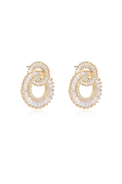 Gold-tone earrings with intricate circular and oval designs are adorned with a mix of clear, rectangular, and round gemstones, reflecting light against a smooth, white background.