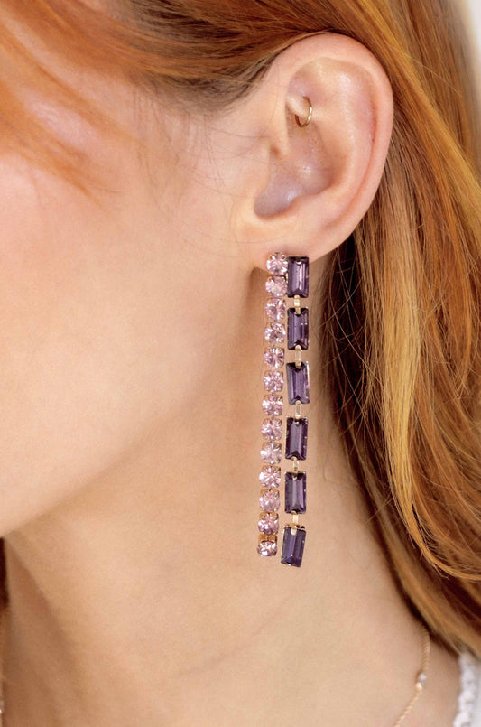 A long, sparkling earring features alternating pink and purple gems, hanging from the ear of a person with reddish-brown hair, set against a neutral background.