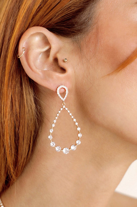 A dangling earring features a teardrop design, shimmering with clear stones. It is worn on a woman's ear, framed by her reddish hair, set against a neutral background.