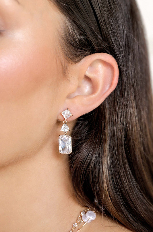 A womanâ€™s ear is adorned with elegant, sparkling drop earrings featuring two large gemstones, one rectangular and one square, set in rose gold, enhancing her side profile against softly flowing hair.