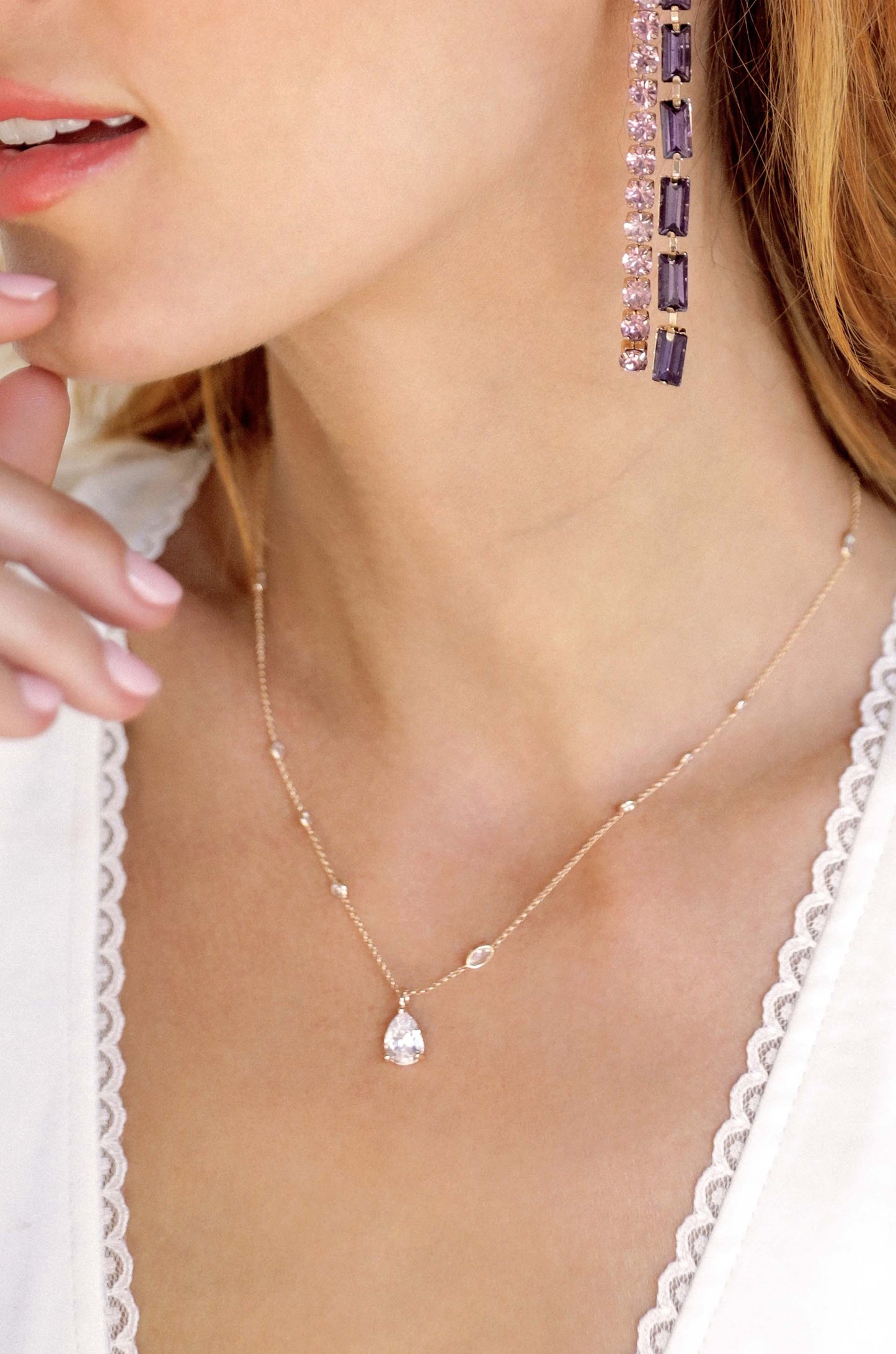 A woman's neck is adorned with a delicate necklace featuring a pear-shaped gemstone, while her ear showcases a long, elegant earring made of purple and pink gemstones. Her hand gently touches her face.