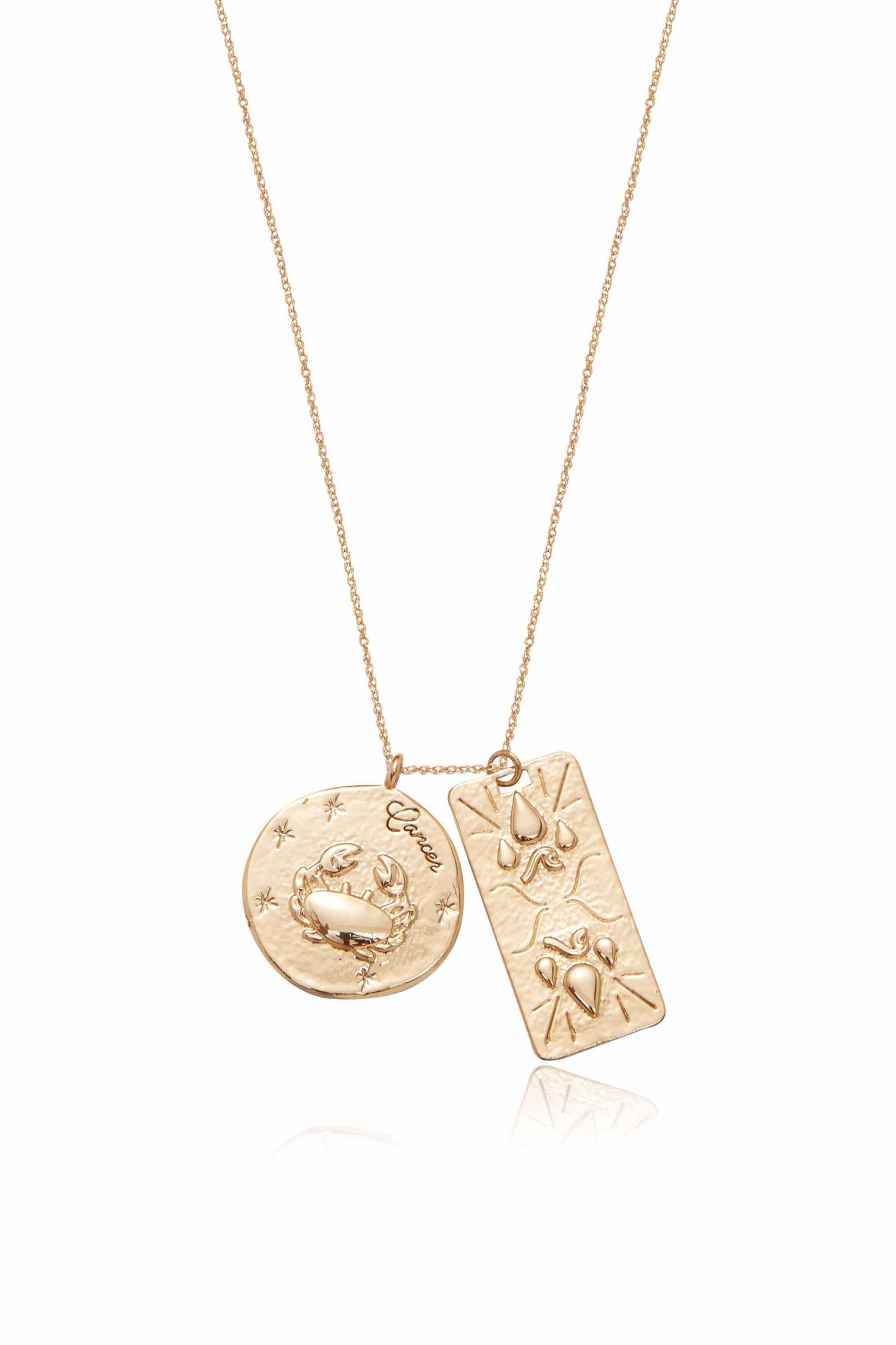 A gold necklace features two pendants: a round one engraved with a crab and the word "Cancer," and a rectangular one with decorative motifs, suspended delicately from a fine chain.