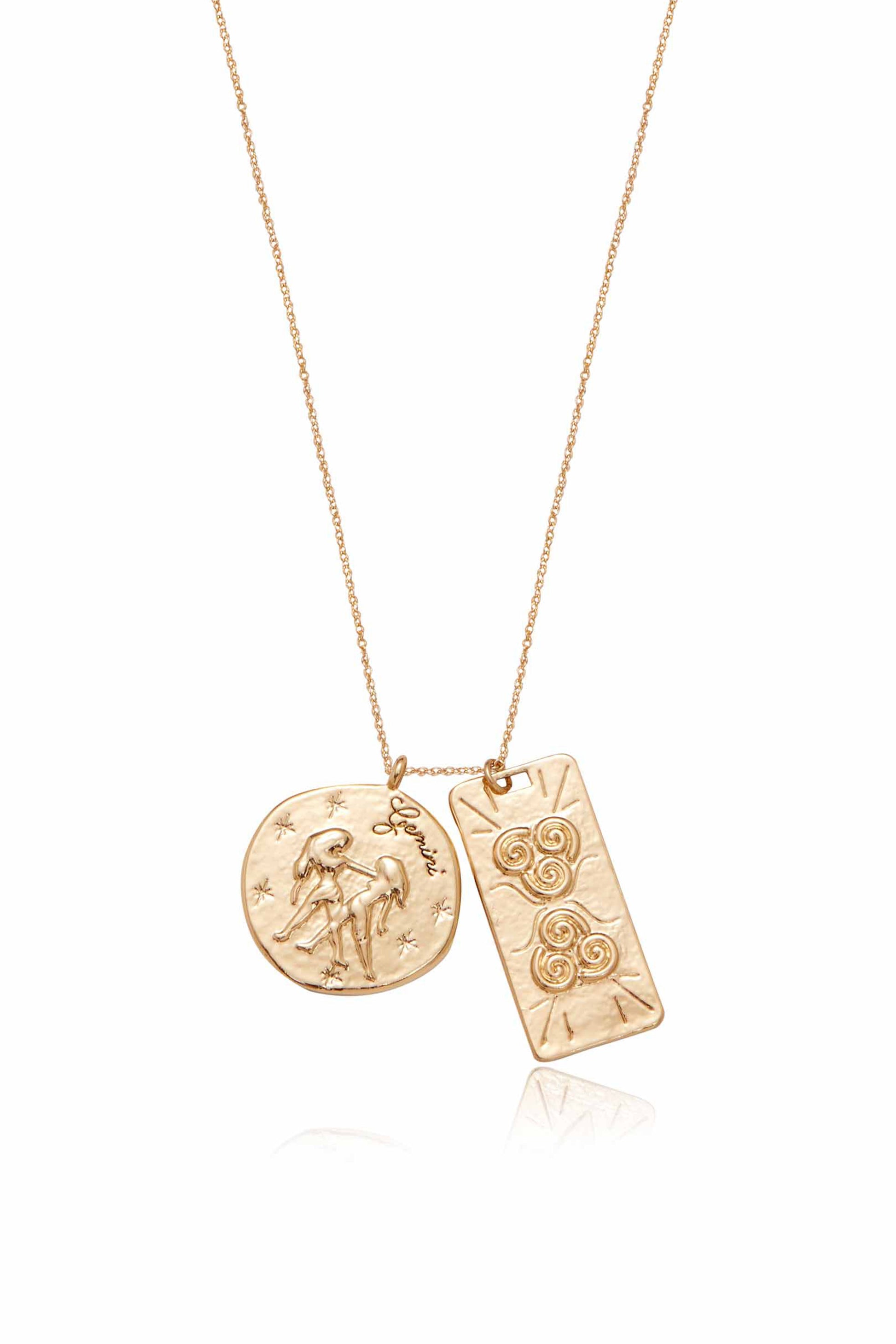 A gold necklace features two pendants: a circular one engraved with Gemini symbols depicting twin figures among stars, and a rectangular pendant with spiral designs. The background is plain white.