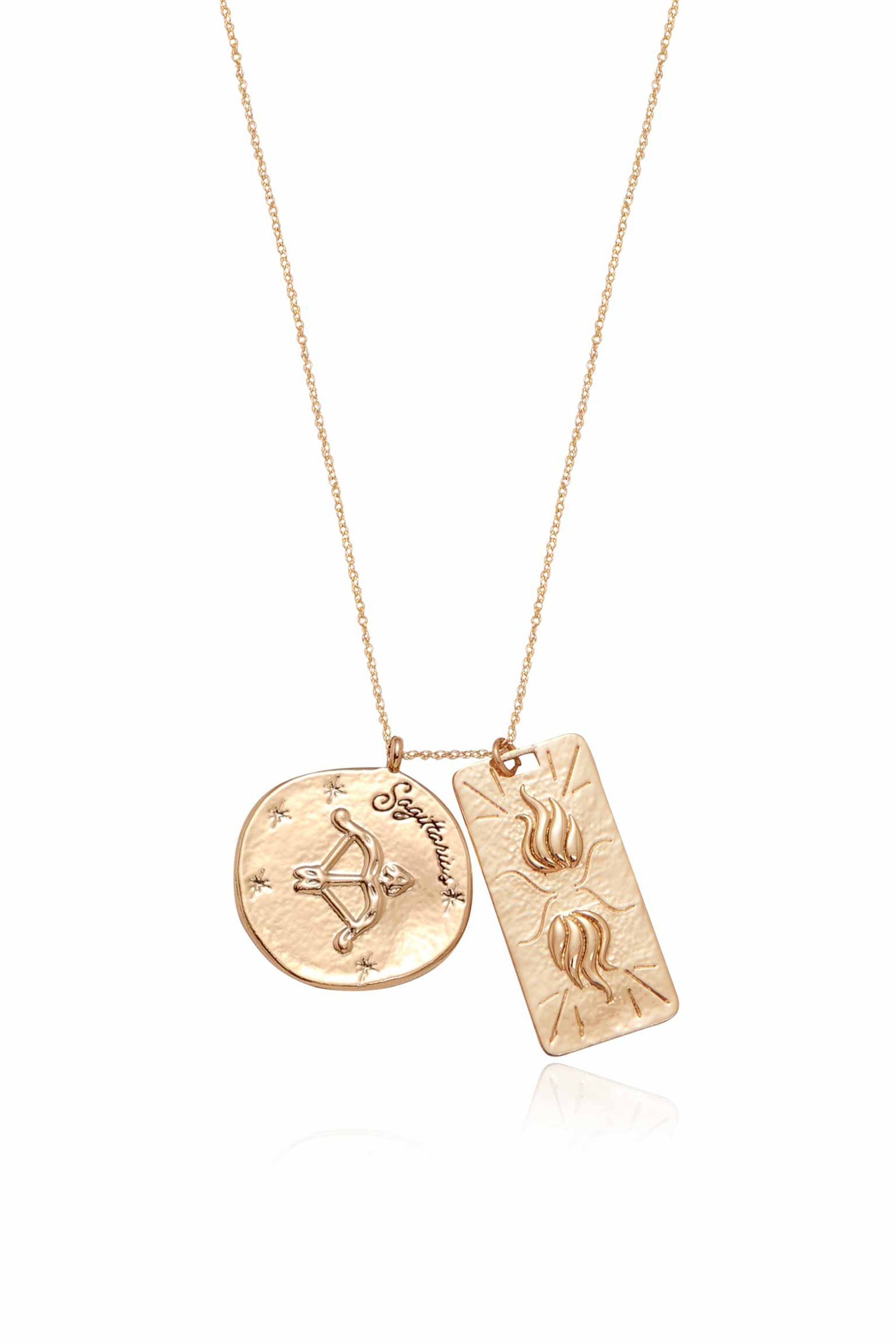 A gold necklace features two pendants: a circular disc with a bow design and "Sagittarius" inscribed, alongside a rectangular pendant with flame motifs, displayed against a reflective surface.