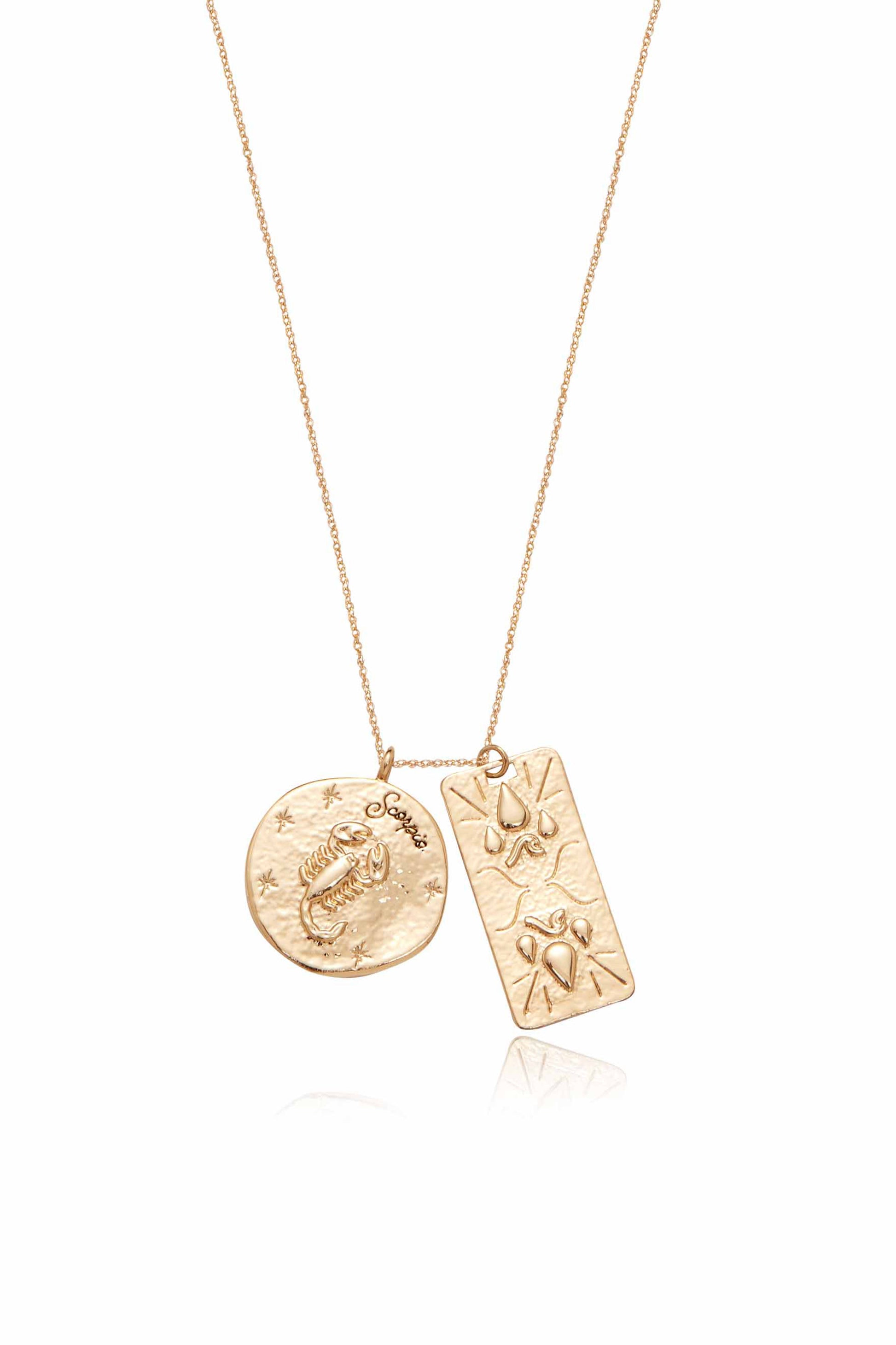 A gold necklace features two pendants: a circular disc with a crab design and the word "Scorpio," alongside a rectangular piece with abstract patterns. The jewelry is displayed against a neutral background.