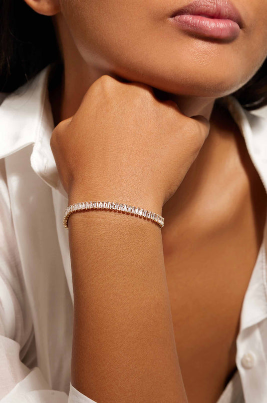 A close-up of a wrist adorned with a sparkling bracelet. The person rests their chin on their hand, showing smooth skin and wearing a white shirt. The background is softly blurred.