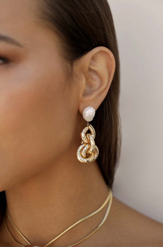 A close-up of an earring featuring a large pearl and an intricate, twisted gold design, hanging from a woman's ear against a neutral background. Subtle jewelry layers complement the elegant accessory.