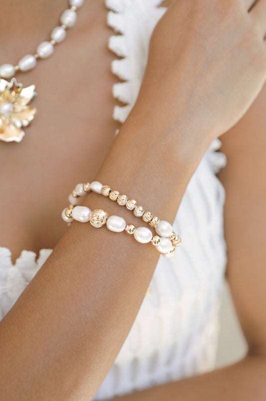 A womanâ€™s arm adorned with two elegant bracelets, featuring a combination of pearls and gold accents. The background is softly blurred, highlighting the jewelry against a light-colored clothing texture.