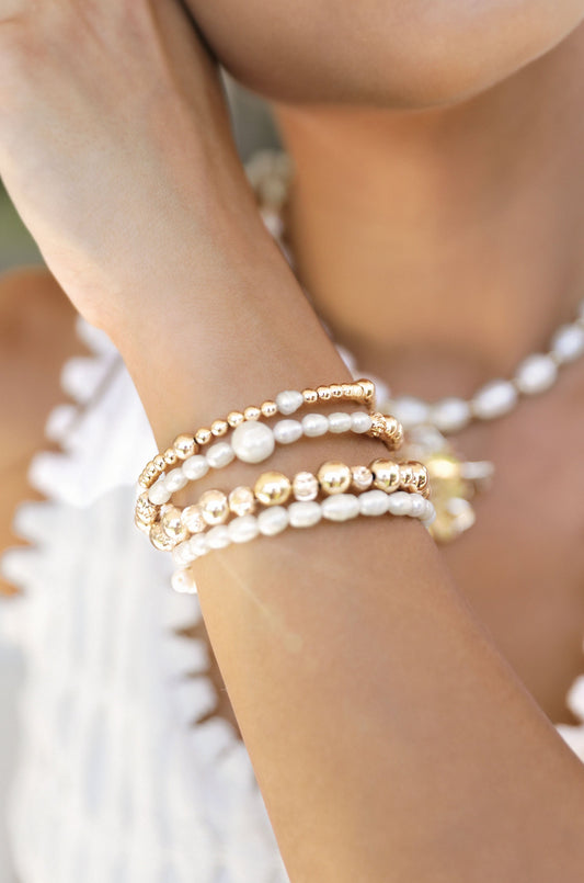 A hand adorned with multiple elegant bracelets made of gold and pearls rests against the skin. The background features soft, blurred colors, suggesting a summery or casual setting.