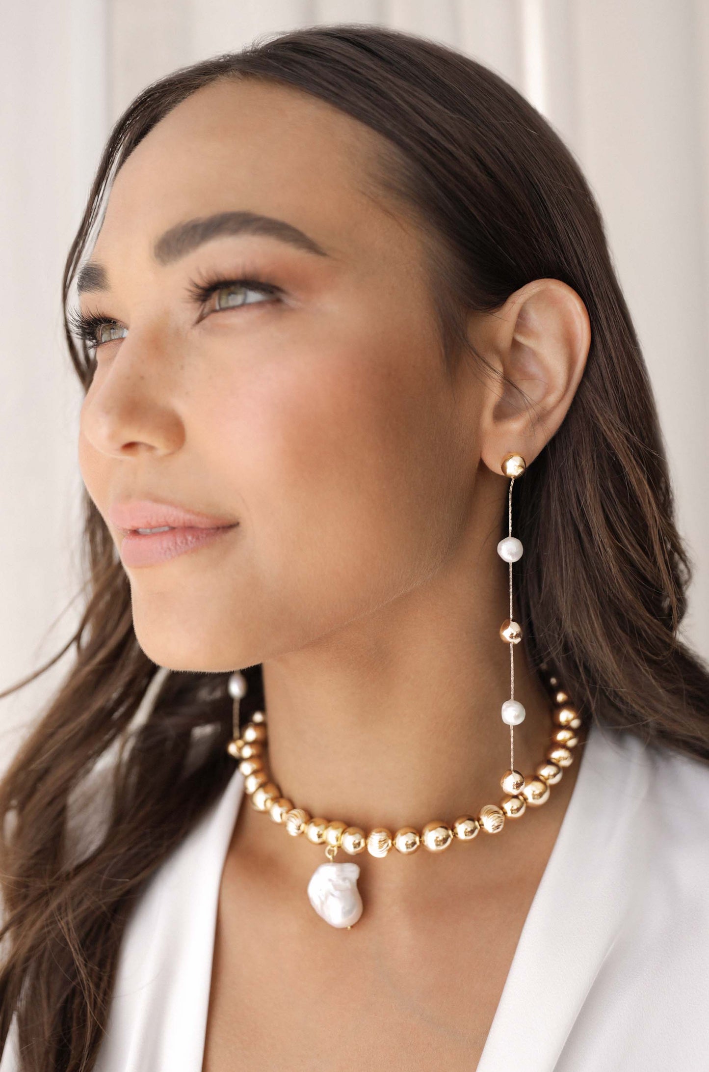 A woman with long, dark hair poses, showcasing a golden bead choker with a pearl pendant and dangling earrings featuring gold and white beads, against a softly lit, neutral backdrop.