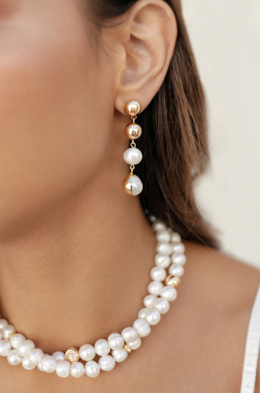 Pearl and gold earrings dangle from the ear, accentuating the neck adorned with a layered pearl necklace. The background is soft and neutral, enhancing the jewelry's elegance.