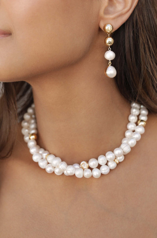 A close-up of a woman's neck, featuring a layered pearl necklace and dangling earrings with pearls. The skin tone is warm, highlighting the jewelry's elegance against a neutral background.