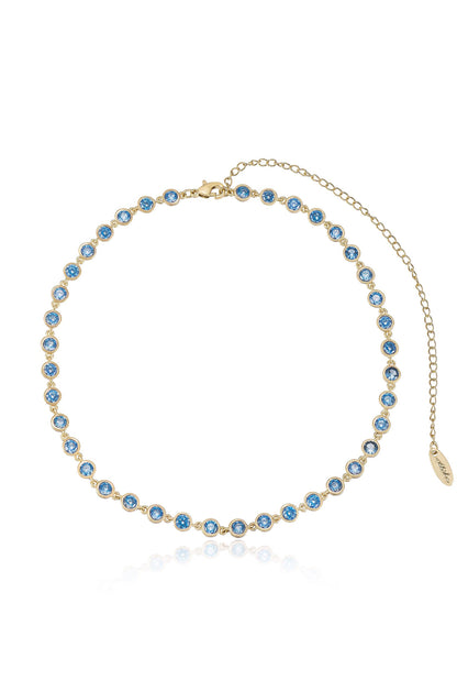 A gold necklace features a continuous line of blue gemstones, each set in a circular bezel. The piece is positioned against a white background, reflecting light off its surface.