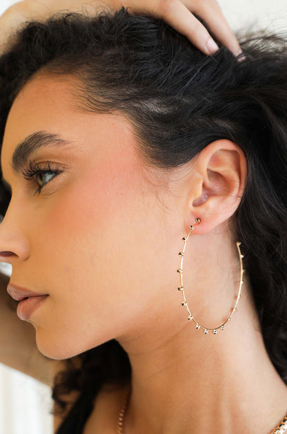 A woman with curly hair touches her head while showcasing a large, thin, gold hoop earring adorned with small decorative elements, against a light-colored background.