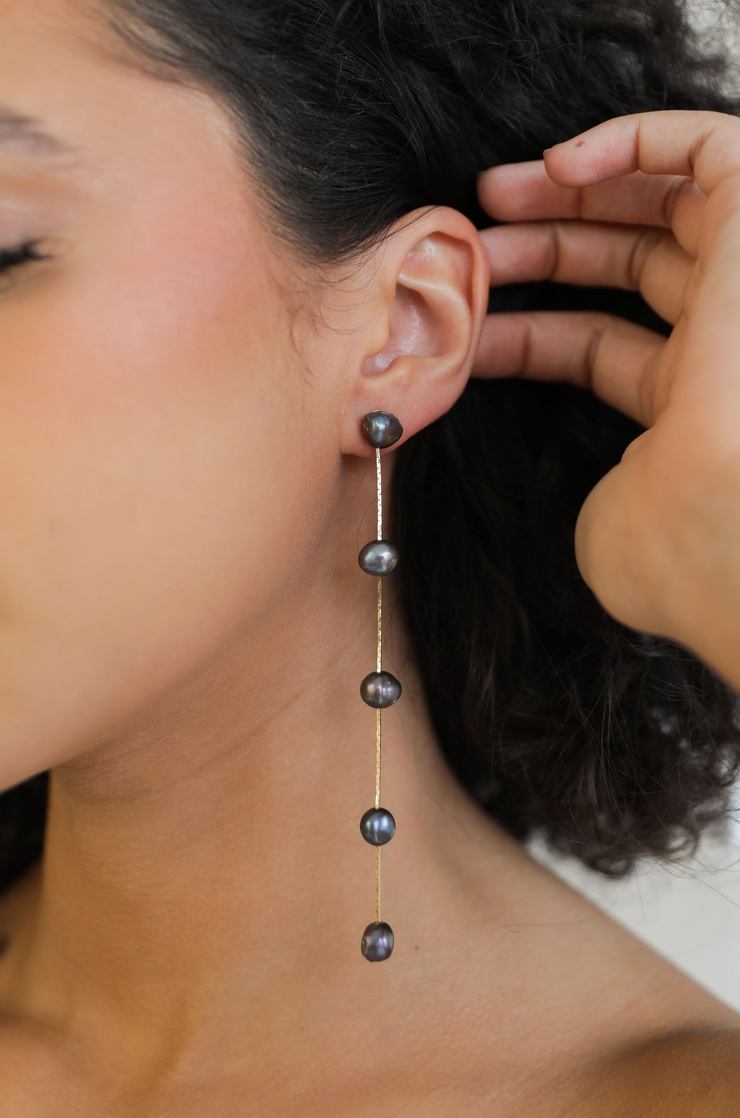 A close-up of an earlobe showcases a dangling earring featuring three dark pearls on a thin gold chain. The person gently touches their hair, with a soft, neutral background.