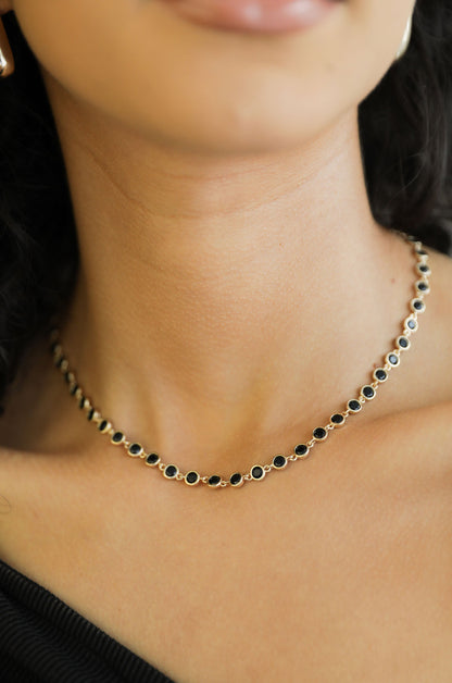 A necklace with black stones in a circular setting rests on a person's neck, accentuating their skin tone. The background is softly out of focus, emphasizing the jewelry.