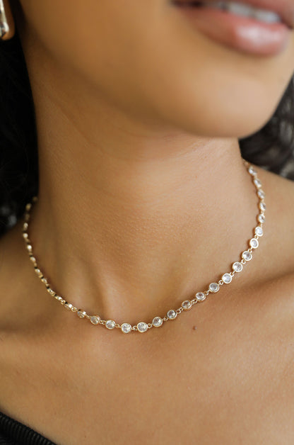 A delicate silver necklace adorned with circular, sparkling stones rests gracefully on a woman's neck, highlighting her smooth skin in a softly lit setting.