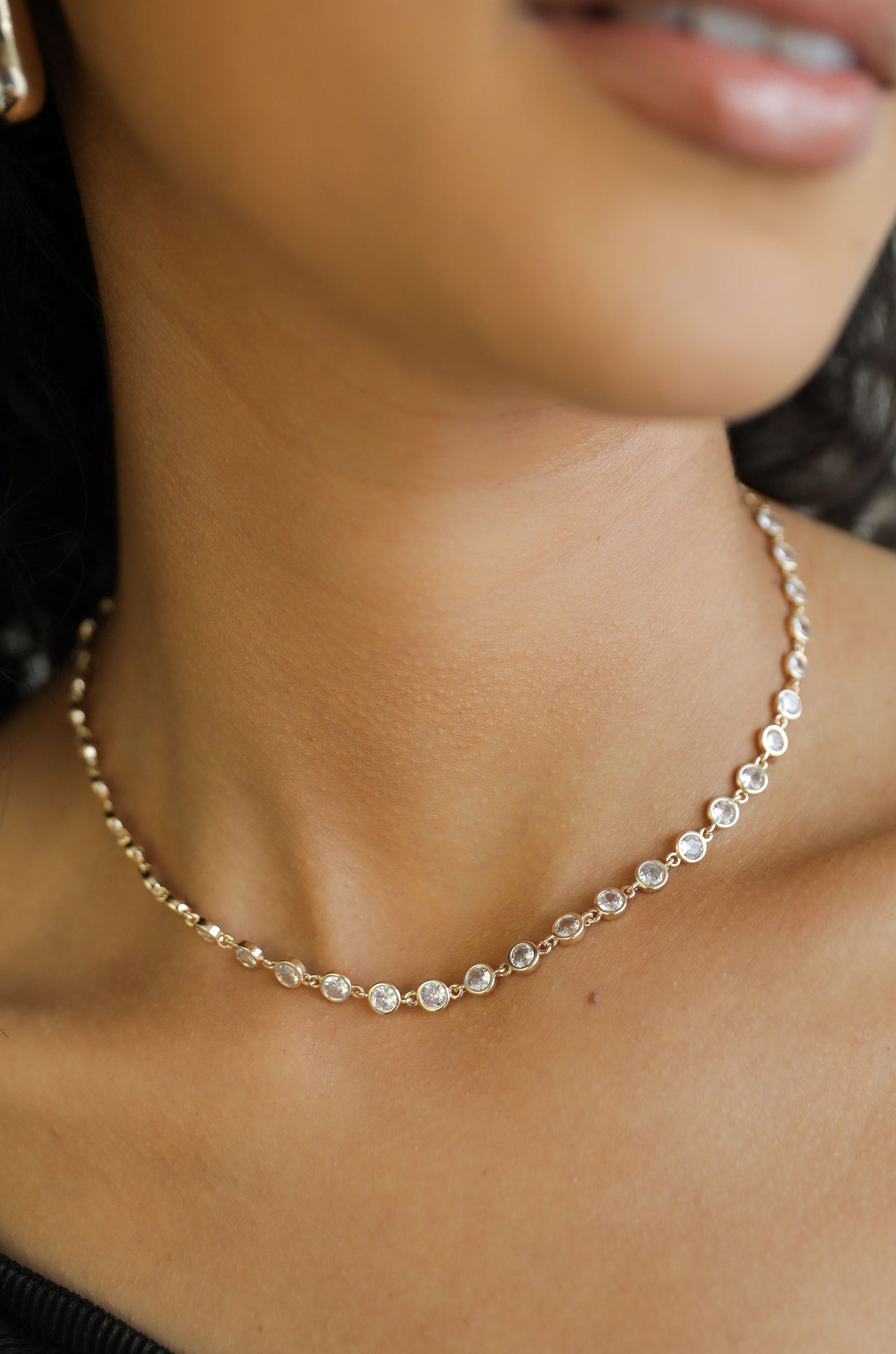 A delicate necklace adorned with circular stones rests on a smooth neck. The individual wears a subtle smile, set against a soft, neutral background, highlighting the elegance of the jewelry.