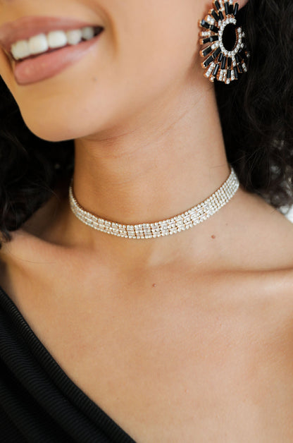 A sparkling choker adorns a woman's neck as she smiles, showcasing her elegant earrings and smooth skin. She wears a dark, textured top, contributing to a sophisticated look.