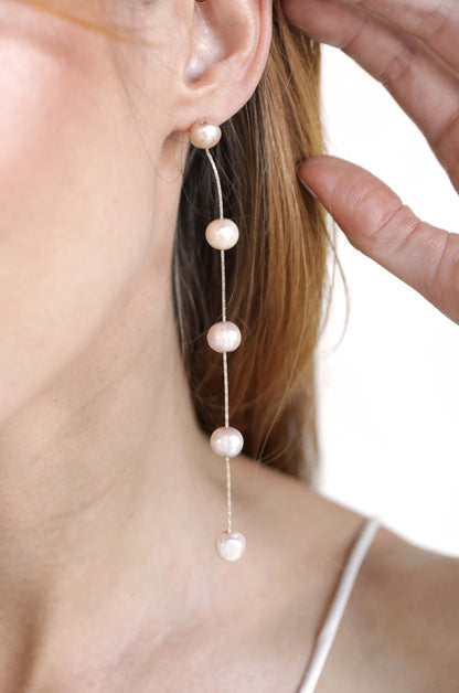 A pair of long, dangling pearl earrings hangs from the ear of a woman, who gently touches her hair, set against a soft, neutral background.