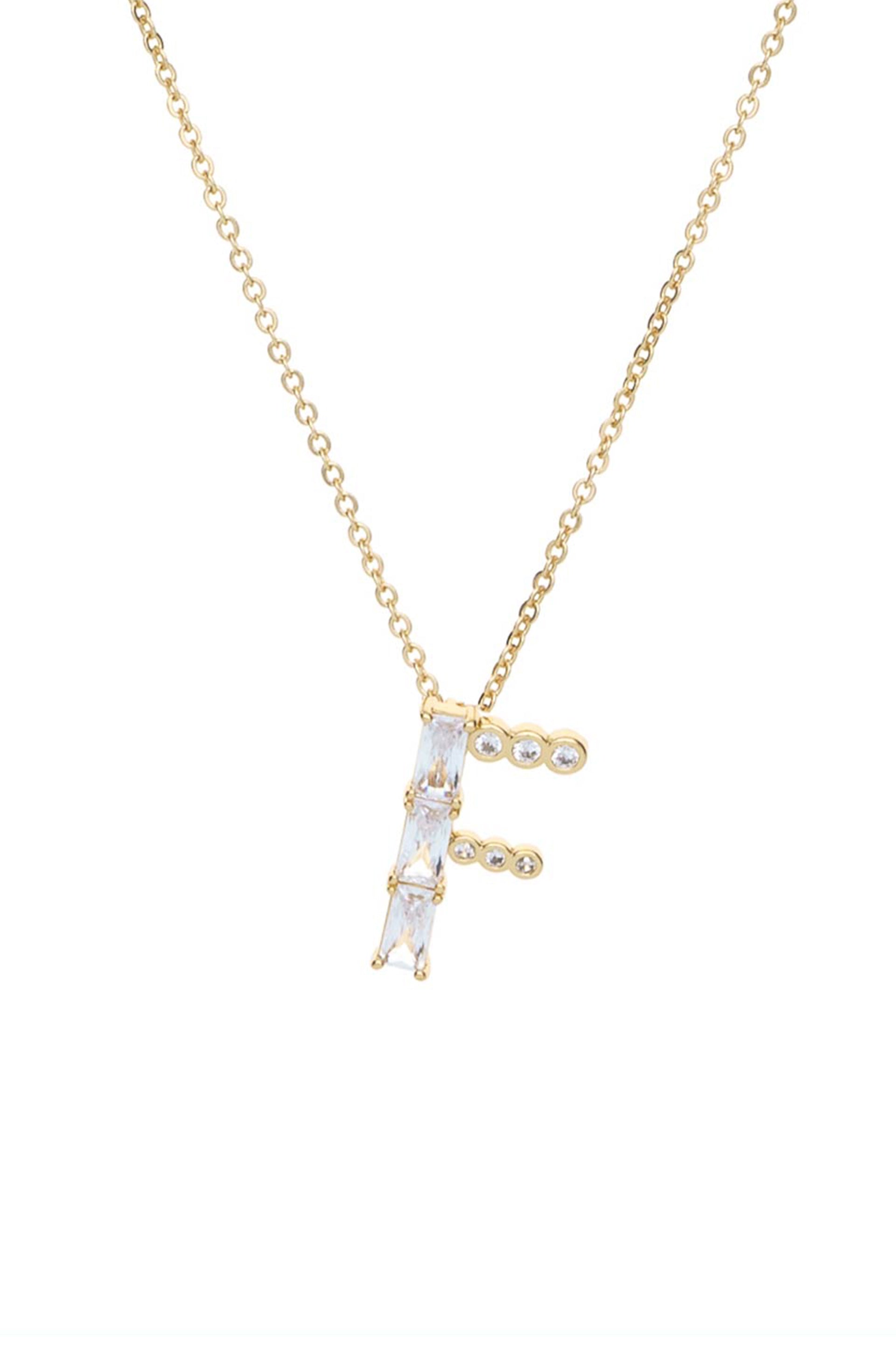 A gold necklace features a decorative letter "F" made of clear, geometric-shaped stones. The pendant hangs from a delicate chain against a plain background, emphasizing its elegance.