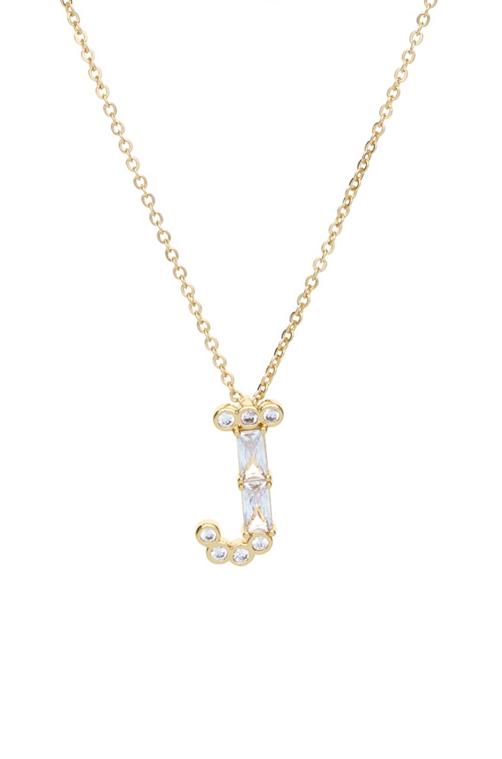 A gold necklace features a letter "J" adorned with clear gemstones, suspended from a delicate chain. The backdrop is plain, emphasizing the jewelry's design and sparkle.