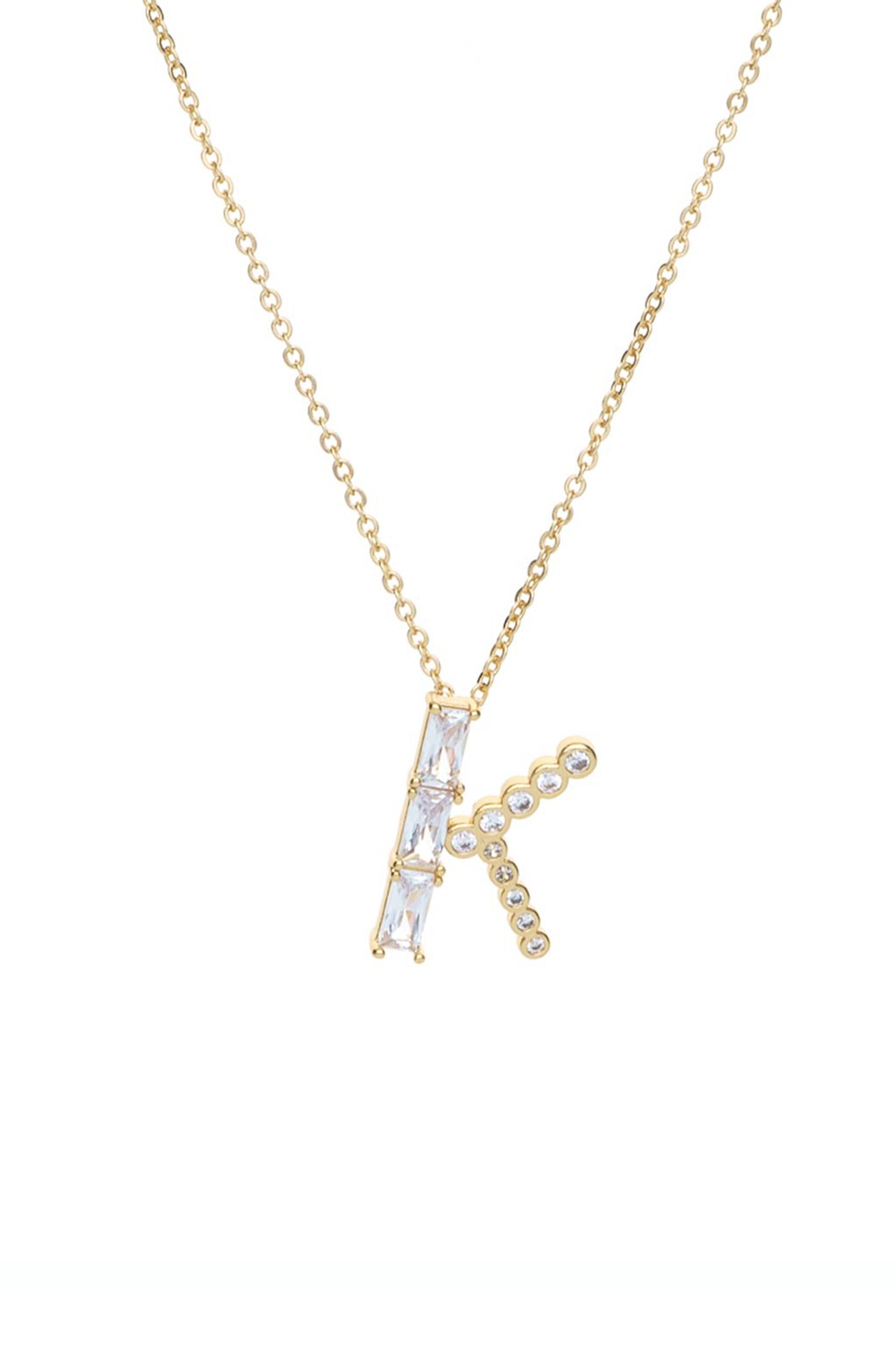 A gold necklace features a stylized letter "K" adorned with clear gemstones. The pendant hangs from a delicate chain, presented on a plain, light background.