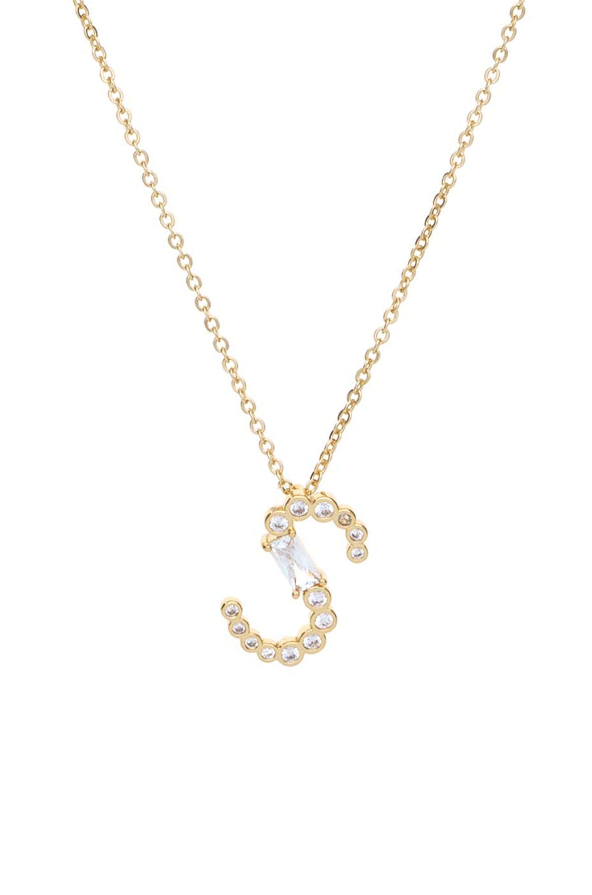 A gold necklace features a stylized "C" pendant adorned with small clear gemstones and a larger diamond-shaped stone, set against a plain white background.