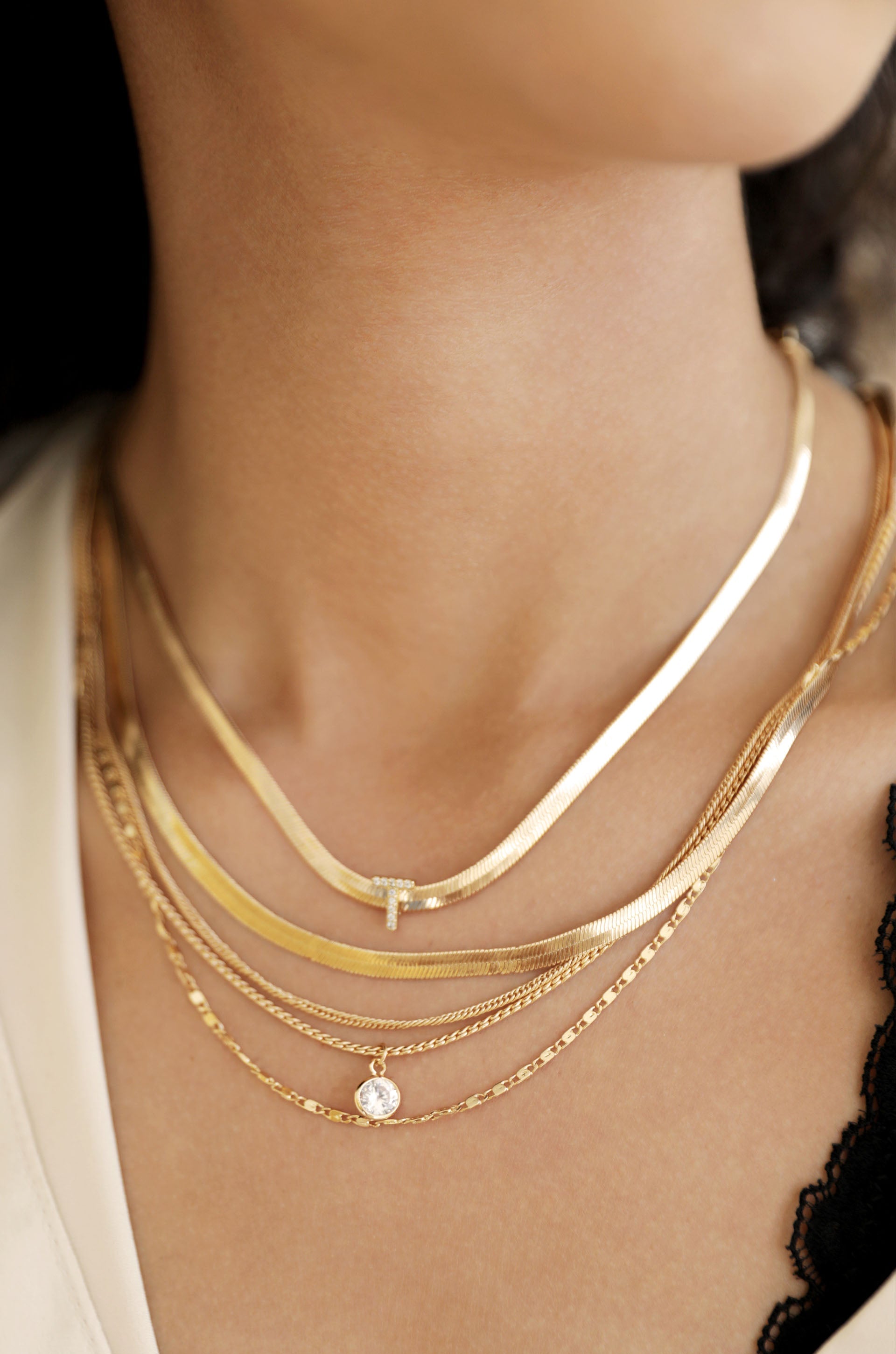 Multiple gold necklaces adorn a person's neck, featuring various designs, including a thin chain with a small pendant. The setting is a softly lit environment, highlighting the jewelry's shine against bare skin.