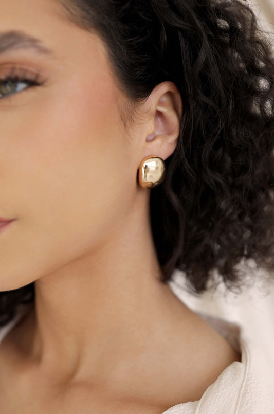 A close-up of a person's ear, adorned with a shiny, gold-colored earring. The individual has curly hair and a natural makeup look, suggesting a relaxed setting.