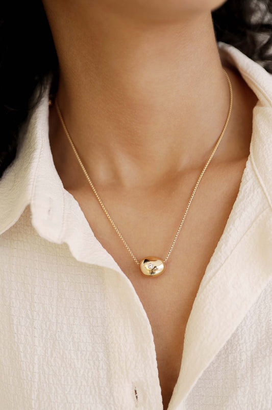 A gold necklace with a smooth, oval pendant featuring a diamond is draped around the neck, resting on the collar of a white, textured shirt.