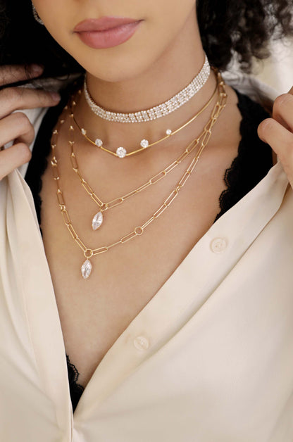 A person is wearing multiple layered necklaces, including a choker and gold chains, against a background of a partially unbuttoned cream blouse, revealing a black lace undershirt.