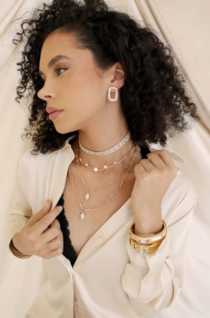 A woman with curly hair poses elegantly, showcasing layered jewelry, including earrings, necklaces, and bracelets. She dons a light-colored shirt over a dark lacy top, against a soft, neutral backdrop.