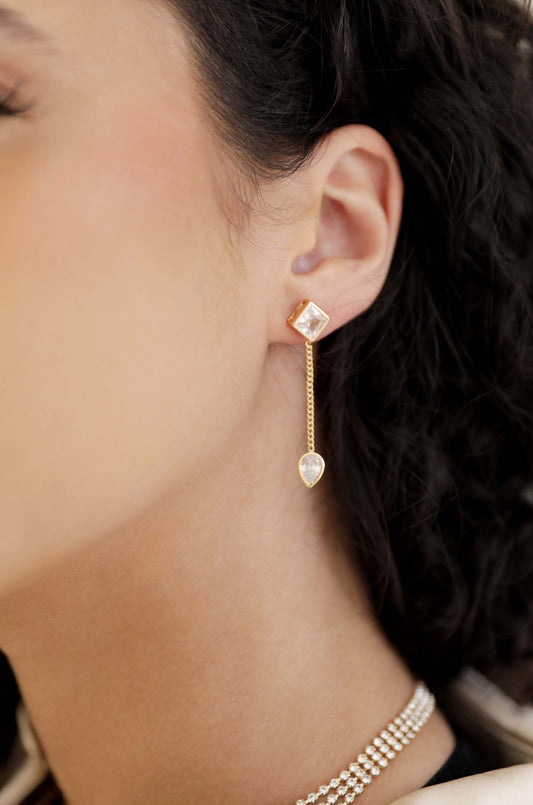 A gold earring featuring a square stud and a dangling teardrop pendant hangs from the model's ear, set against her smooth skin and curly hair, showcasing a stylish accessory.