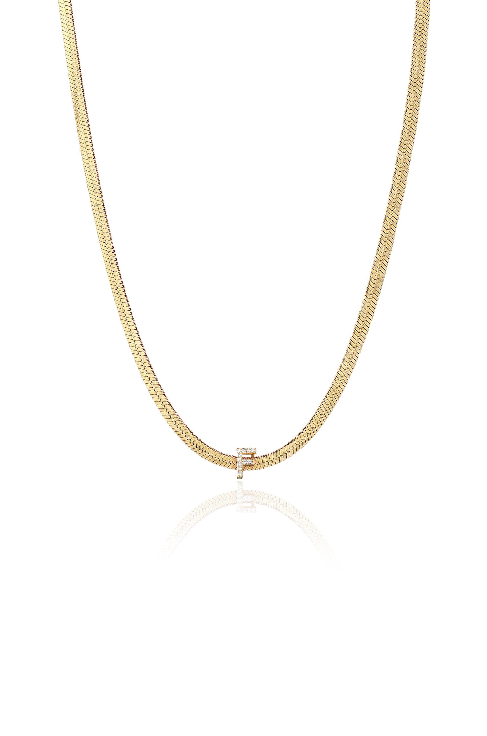 A gold chain necklace displays a sleek design with a small, polished letter "E" pendant. The necklace is reflected on a smooth, light-colored surface, enhancing its elegance.