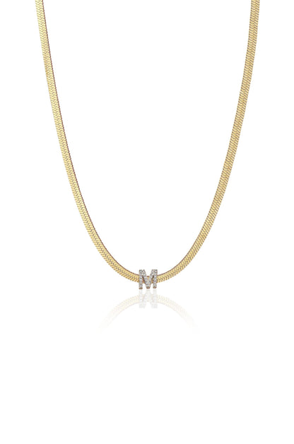 A gold necklace features a smooth herringbone design. A sparkling diamond-studded letter "M" pendant hangs centrally, reflecting light subtly on a clean, white background.