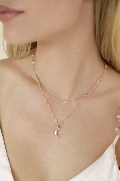 A delicate layered gold necklace with a crescent moon pendant rests against a bare neck, surrounded by soft, neutral skin tones and the elegant drape of a light garment.