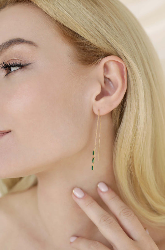 A woman gently touches her neck while showcasing a long, delicate gold earring with three green stones, set against a softly illuminated background that emphasizes her features and jewelry.