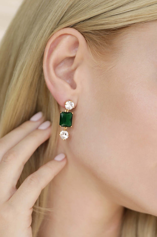 A hand gently touches a large green gemstone earring with two sparkling diamonds attached, set against a close-up of a woman's ear and stylish, straight blonde hair.