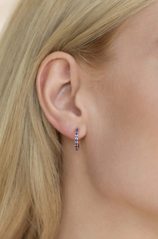 A close-up of a womanâ€™s ear featuring a gold hoop earring adorned with blue gemstones. The background is softly blurred, emphasizing the earring and the woman's smooth, fair skin.