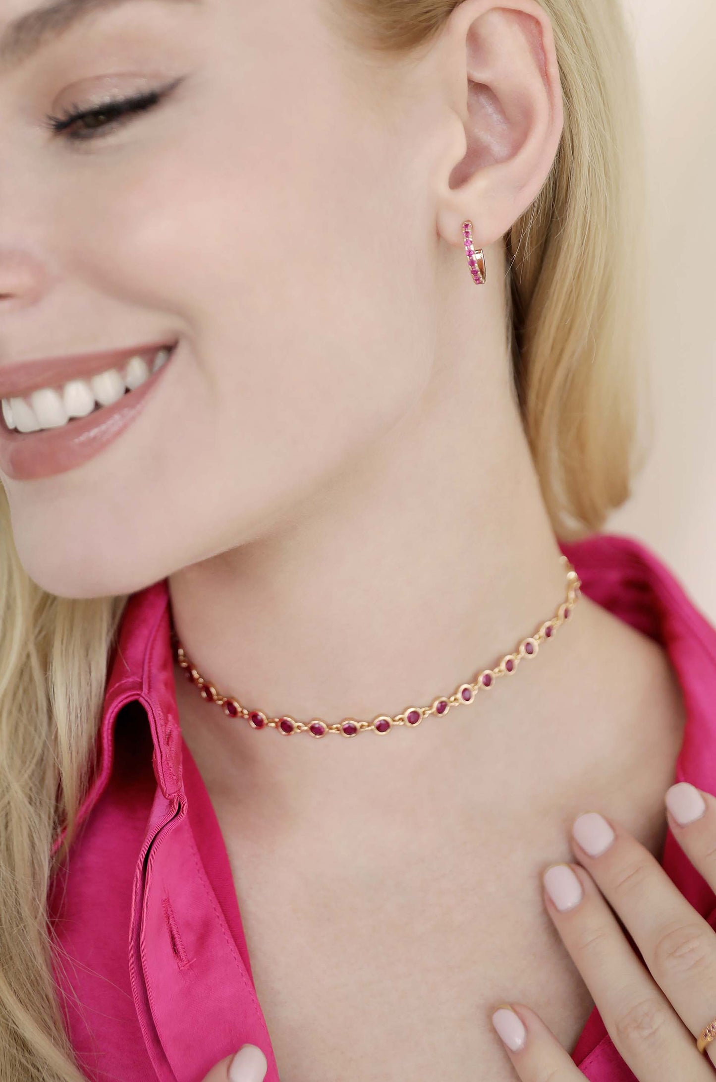 A woman wears a pink choker and matching drop earrings, smiling softly. She's dressed in a pink shirt, with her hair cascading nearby, set against a light, neutral background.