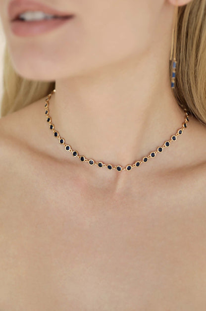 A gold necklace with circular black stones is worn around a delicate neck. The background is softly blurred, highlighting the jewelry and the subtle features of the person's collarbone.